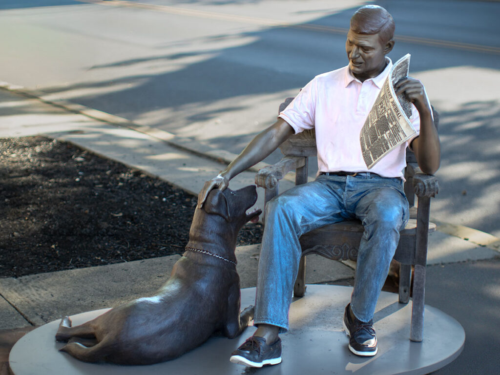A bronze sculpture titled "Keep in Touch" depicts a man reading a newspaper and reaching down affectionately for the dog at his feet.